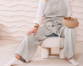May include: A person wearing a light gray linen apron with a brown leather belt and a white shirt, sitting on a white stool. The person is holding a woven basket.