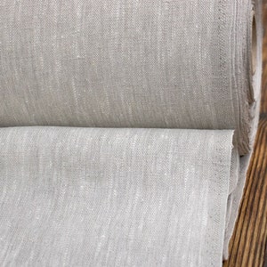 Op de afbeelding: Een rol natuurlijk gekleurde linnen stof. De stof heeft een getextureerde uitstraling en een neutrale, lichtbeige tint. De rol is gedeeltelijk afgerold, waardoor de weefsel en dikte van de stof zichtbaar zijn. De achtergrond is een donker houten oppervlak.