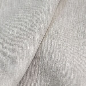 Op de afbeelding: Close-up van lichtgrijze linnen stof. De stof is gevouwen en toont de textuur en weefsel. Het materiaal lijkt zacht en heeft een natuurlijk, licht ongelijkmatig uiterlijk, geschikt voor kleding of interieurdecoratie.
