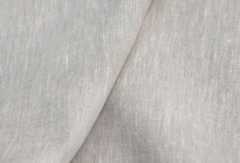 Op de afbeelding: Close-up van lichtgrijze linnen stof. De stof is gevouwen en toont de textuur en weefsel. Het materiaal lijkt zacht en heeft een natuurlijk, licht ongelijkmatig uiterlijk, geschikt voor kleding of interieurdecoratie.