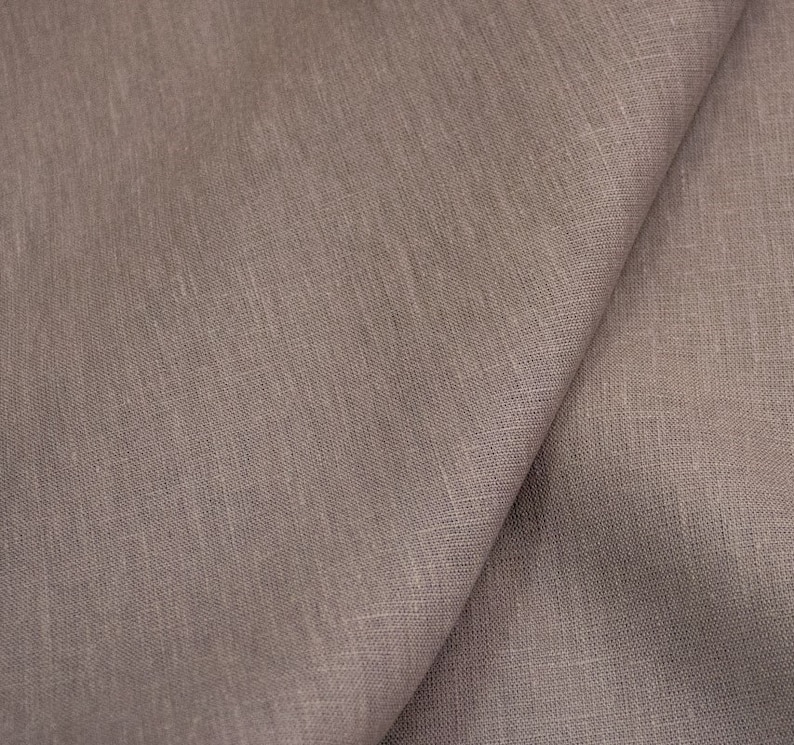 Op de afbeelding: Close-up van een gevouwen stuk taupekleurig linnen. De stof heeft een getextureerde uitstraling met zichtbare weefdetails. De plooien cre&euml;ren schaduwen, die de drapering en textuur van het materiaal benadrukken. De algehele esthetiek is eenvoudig en natuurlijk.