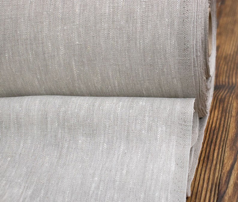 Op de afbeelding: Een rol natuurlijk gekleurde linnen stof. De stof heeft een getextureerde uitstraling en een neutrale, lichtbeige tint. De rol is gedeeltelijk afgerold, waardoor de weefsel en dikte van de stof zichtbaar zijn. De achtergrond is een donker houten oppervlak.