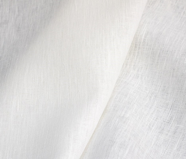 Op de afbeelding: Close-up van witte linnen stof met een getextureerde weefsel. De stof is gevouwen, waardoor schaduwen ontstaan en de natuurlijke textuur en kleurvariaties worden benadrukt. De afbeelding toont de zachte, ademende kwaliteit van de stof.