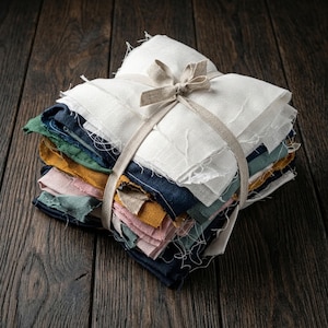 May include: A stack of square fabric swatches with frayed edges, tied together with a light beige ribbon. The fabric pieces are in various colors including white, dark blue, green, mustard yellow, and soft pink, layered in a neat bundle.