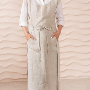 Puede incluir: Un delantal largo de lino con un lazo en la cintura y dos bolsillos grandes. El delantal es de color beige claro.