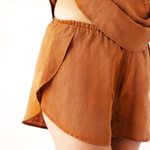 May include: Close-up of a pair of brown linen shorts with a curved hemline. The shorts have a drawstring waistband and are made of a soft, lightweight fabric.