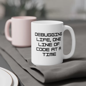 Peut inclure: Mug en céramique blanche avec le texte "DEBUGGING LIFE, ONE LINE OF CODE AT A TIME" en noir. Un deuxième mug rose est légèrement flou en arrière-plan. Les mugs sont sur une surface grise.
