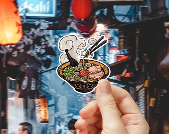 May include: A cartoon sticker of a bowl of ramen with chopsticks, steam, and a cute face. The sticker is held in a hand against a blurred background of a Japanese street.