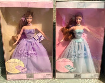 Op de afbeelding: Twee Barbie-poppen in dozen. De ene pop draagt een lavendelkleurige jurk, de andere een lichtblauwe jurk. Beide poppen hebben lang haar en zitten in dozen met de tekst "Birthday Wishes". Elke doos bevat een klein cadeautje en het Barbie-logo.