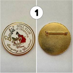 Op de afbeelding: Witte en gouden pin met een rood en wit beeld van een meisje in een slee met de tekst "Barbie Doll Club of Oklahoma Where Everyday is Christmas". De achterkant van de pin is goud met een pin.