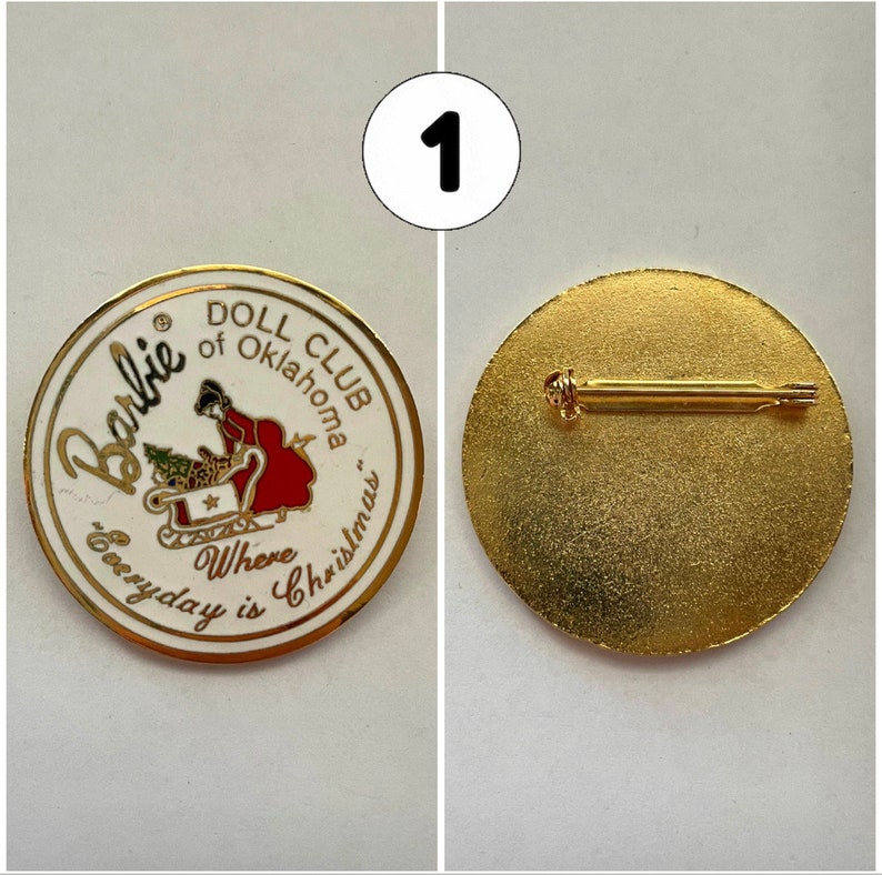 Op de afbeelding: Witte en gouden pin met een rood en wit beeld van een meisje in een slee met de tekst "Barbie Doll Club of Oklahoma Where Everyday is Christmas". De achterkant van de pin is goud met een pin.