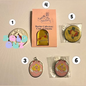 Op de afbeelding: Een verzameling van tien Barbie-verzamelaarspinnen. De pinnen zijn in verschillende kleuren en hebben verschillende ontwerpen, waaronder een pop, een hart en een trouwjurk. De pinnen zijn genummerd van 1 tot en met 6.
