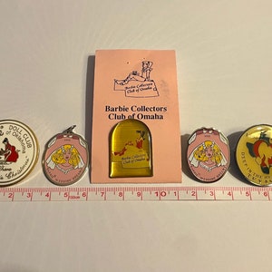 Op de afbeelding: Vijf verschillende Barbie-verzamelpinnen. De pinnen zijn allemaal rond en hebben verschillende ontwerpen. E&eacute;n pin heeft een kerstsc&egrave;ne met een rode slee en een kerstboom. Een andere pin heeft een huwelijkssc&egrave;ne met een bruid in een witte jurk. Een derde pin heeft een kaart van Omaha, Nebraska. Een vierde pin heeft een bruid in een witte jurk met de tekst "Barbie's Wedding Dream". De vijfde pin heeft een bruid in een witte jurk met de tekst "Deep in the Heart of Texas".