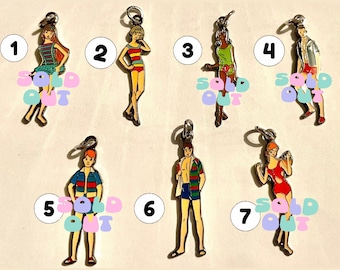 Op de afbeelding: Zeven kleurrijke emaille hangers met mensen in verschillende zomeroutfits. De hangers zijn genummerd van 1 tot 7 en hebben een 'uitverkocht' label.