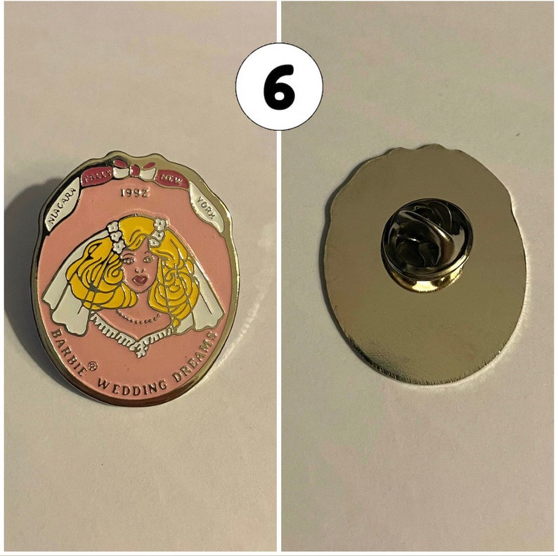 Op de afbeelding: Een roze en witte emaille pin met een cartoonbruid met blond haar en een witte sluier. De pin heeft de tekst "Barbie Wedding Dreams" en "1992" aan de voorkant. De achterkant van de pin is zilver met een pin achterkant.