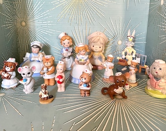 May include: A collection of 14 vintage porcelain figurines of nurses, including a bunny rabbit, a dog, and a mouse. The figurines are all wearing white uniforms with red crosses and are holding various medical supplies.