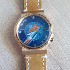May include: Vintage gold-coloured wristwatch with a blue face featuring a rocket and globe design. The watch face includes the text "CCCP" and Cyrillic lettering. It has a tan leather strap with white stitching.