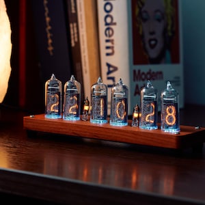 Pode incluir: Um relógio de tubos nixie com seis tubos de vácuo de vidro montados numa base retangular de madeira. Os tubos exibem dígitos laranja brilhantes, com dois tubos mais pequenos entre os maiores, emitindo uma luz azul suave.