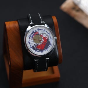 May include: A silver-cased wristwatch with a black leather band and white stitching. The watch face features a map design with red and blue accents. The watch is displayed on a wooden and black leather stand.