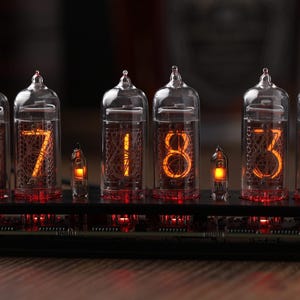 Könnte beinhalten: Eine Digitaluhr mit sechs Glasröhren, die die Uhrzeit 17:34 anzeigt. Die Ziffern leuchten orangefarben vor dunklem Hintergrund. Die Uhr steht auf einem schwarzen Sockel mit sichtbaren elektronischen Bauteilen.