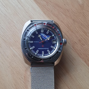 Op de afbeelding: Roestvrijstalen polshorloge met een blauwe wijzerplaat met witte markeringen en wijzers, een rode ster en de tekst "Автоматический". Het horloge heeft een mesh band en een datumweergave op 3 uur.