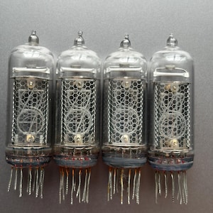 Può includere: Quattro tubi nixie in vetro trasparente disposti in fila, ciascuno caratterizzato da un bulbo a vuoto cilindrico con griglie in rete metallica interna e numeri in filo metallico impilati. Ogni tubo ha una punta in vetro e dei pin metallici.
