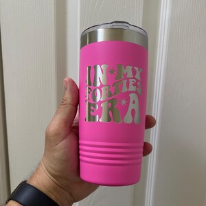 Könnte beinhalten: Ein leuchtend pinkfarbener Isolierbecher mit silbernem Deckel. Der Becher trägt den Text "IN MY FORTIES ERA" in einem Retro-Schriftstil. Der zylindrische Becher hat horizontale Rillen.