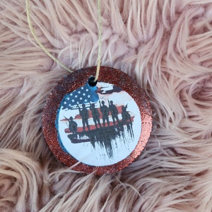 May include: A round, red glitter ornament with a white background featuring a silhouette of five soldiers standing in front of an American flag.