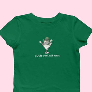 May include: A green t-shirt with a cartoon illustration of a leprechaun in a martini glass. The text "drinks well with others" is printed below the image.