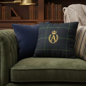 May include: A collection of decorative throw pillows on a green sofa. One pillow is navy blue, and another is a dark green plaid with a gold embroidered crown and the letter "A". The sofa is in front of a bookshelf.