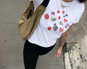 Puede incluir: Una camiseta blanca con un estampado de frutas coloridas que incluye fresas, cerezas, arándanos y moras. La camiseta se lleva con pantalones negros y un bolso marrón.