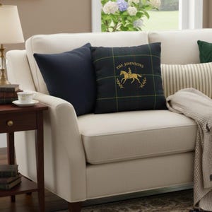 May include: A cream sofa with two cushions. One is navy, the other a dark green plaid with a gold equestrian design and the text "THE JOHNSONS". A beige throw blanket is draped over the arm. The sofa is in front of a window.