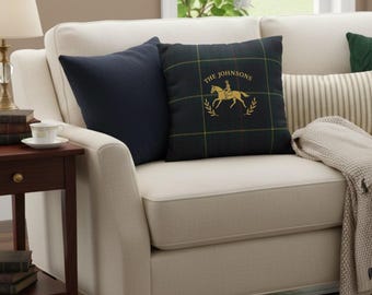 May include: A cream-colored sofa with two throw pillows. One pillow is navy blue, and the other is a dark green plaid with a gold equestrian design and the text "THE JOHNSONS". A beige throw blanket is draped over the arm.