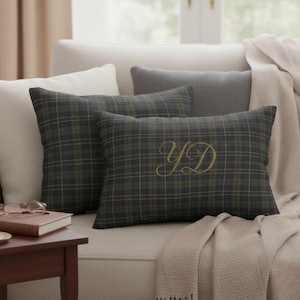 May include: Two decorative pillows with a green and navy plaid pattern and gold embroidered initials on a cream-colored sofa. A gray pillow and a cream-colored throw blanket are also visible. A small wooden side table with a book and glasses is in the foreground.