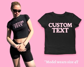 May include: Black cropped t-shirt with pink "CUSTOM TEXT" lettering. The model is wearing size 4T.