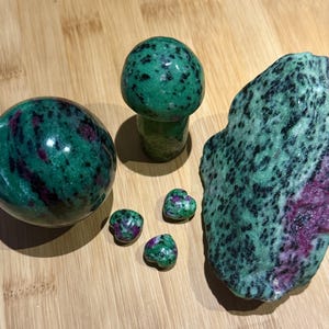 May include: A collection of polished zoisite stones with ruby inclusions. The arrangement includes a sphere, a mushroom-shaped carving, a large, rough-cut piece, and several heart-shaped stones. The stones display a mix of green and red hues.