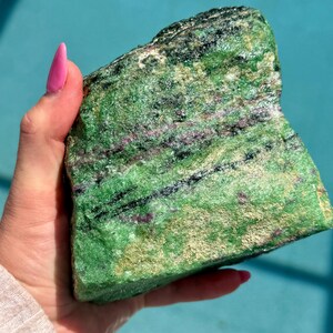 May include: A rough-cut, natural green and black stone with streaks of red and gold. The stone is held in a hand with pink nail polish, against a turquoise background. The stone's texture is rough and uneven, with visible mineral deposits.