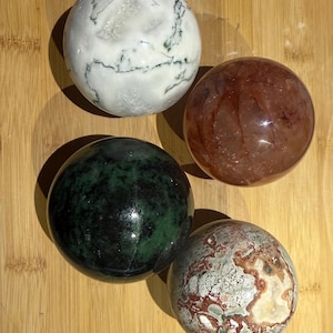 May include: Four polished gemstone spheres in varying colors. One is white with green veining, another is a deep red-brown, a third is a dark green, and the last is a mix of beige, red, and gray patterns. The spheres are displayed on a light brown wooden surface.