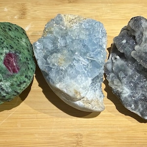 May include: Three raw mineral specimens: a green zoisite with ruby inclusions, a light blue celestite cluster, and a dark gray druzy quartz. The minerals are displayed on a wooden surface.