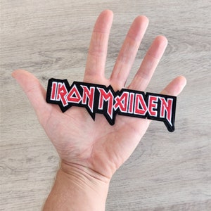 Può includere: Una toppa ricamata nera e rossa con la scritta "IRON MAIDEN" in un font stilizzato. La toppa è rettangolare con un bordo frastagliato ed è tenuta in una mano.