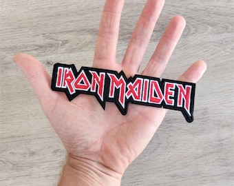 May include: A black and red embroidered patch featuring the words "IRON MAIDEN" in a distinctive font. The patch has a jagged edge and is held in a hand.