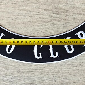 May include: Black, curved patch with the text "NO CLUB" in white, with a white border. A yellow tape measure shows the patch is approximately 14 inches long. The patch is on a light brown wooden surface.