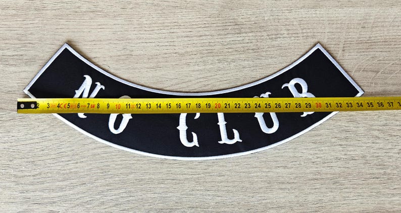 May include: Black, curved patch with the text "NO CLUB" in white, with a white border. A yellow tape measure shows the patch is approximately 14 inches long. The patch is on a light brown wooden surface.