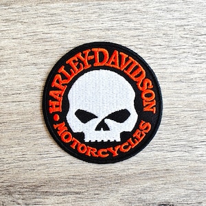Peut inclure: Un écusson brodé noir et orange avec un crâne et des os croisés blancs au centre. Le texte "Harley-Davidson Motorcycles" est écrit en cercle autour du crâne.