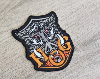 May include: Embroidered patch depicting a stylized boar's head with red eyes, white tusks, and grey and white fur. The patch has an orange and black border with the number "10" and a circular design.
