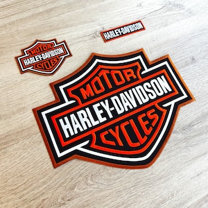 Può includere: Un grande patch ricamato arancione e nero con le parole "MOTOR", "HARLEY-DAVIDSON" e "CYCLES" a forma di scudo.
