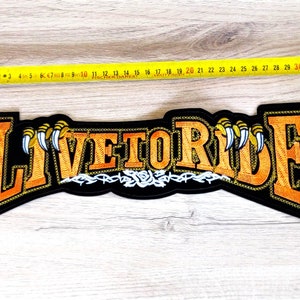 Può includere: Una toppa ricamata nera con le parole "LIVE TO RIDE" in arancione con artigli bianchi su entrambi i lati. La toppa è larga circa 28 pollici.