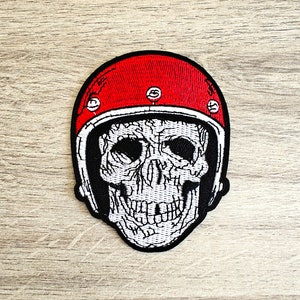 Pode incluir: Um remendo bordado em preto e branco de uma caveira usando um capacete de moto vermelho. O capacete tem detalhes brancos e cinco rebites prateados.