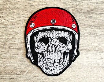 May include: A black and white embroidered patch of a skull wearing a red motorcycle helmet. The helmet has white accents and five silver rivets.