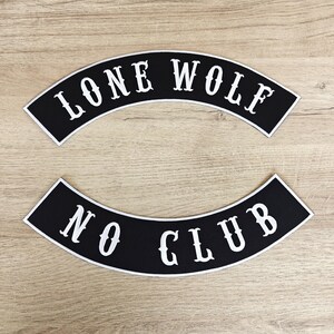 May include: Two black, arched patches with white text. The top patch reads "LONE WOLF" and the bottom patch reads "NO CLUB". The patches have a white border and are set against a light wood grain background.